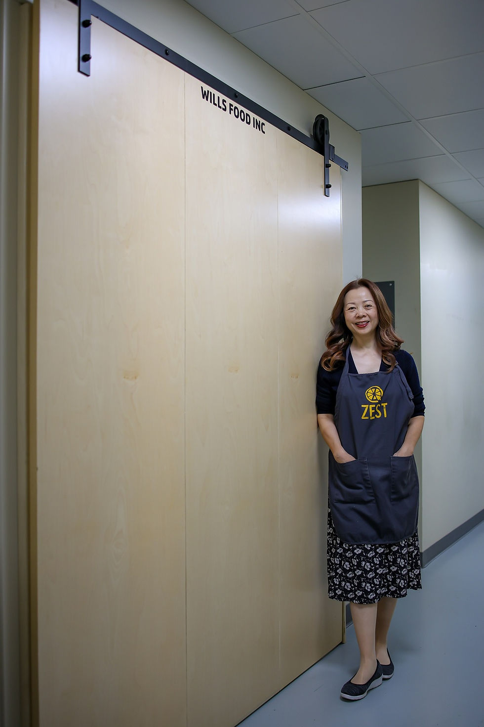 Woman in a Zest apron smiles, standing in a hallway by a light wooden door marked "Wills Food Inc." Gray floor and neutral walls.