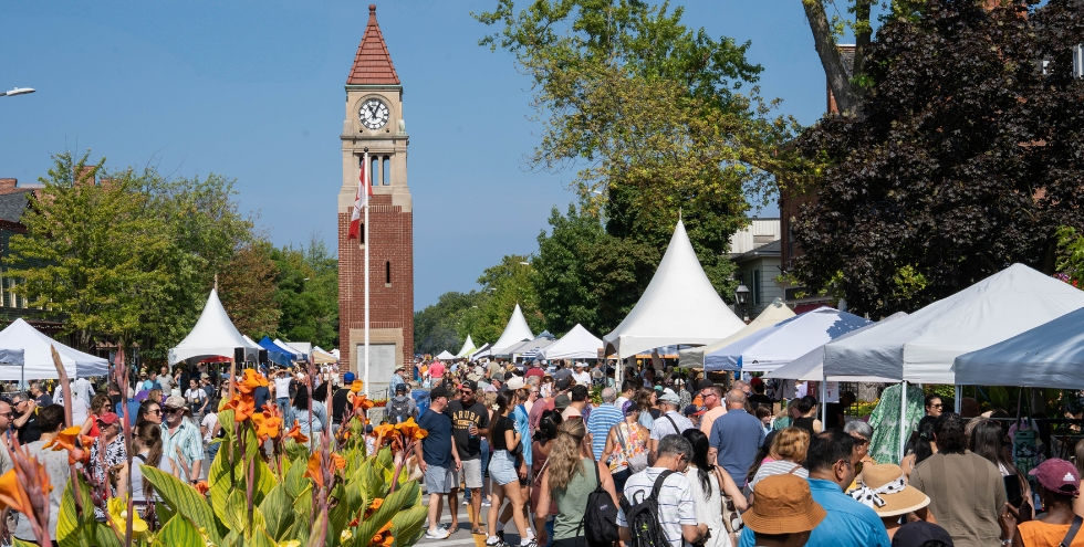 The Peach Festival on Queen St in Niagara-on-the-Lake. Outdoor market with white tents and a clock tower, people browsing under a sunny blue sky, flowers in foreground.
