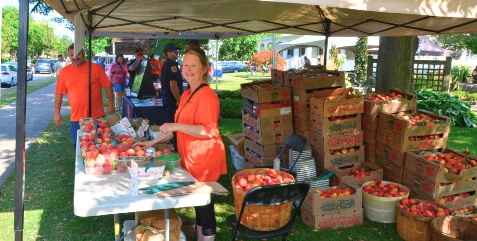 Peach Festival in Niagara-on-the-Lake. Smiling woman in orange sells peaches under a canopy, with stacked boxes and baskets of peaches.