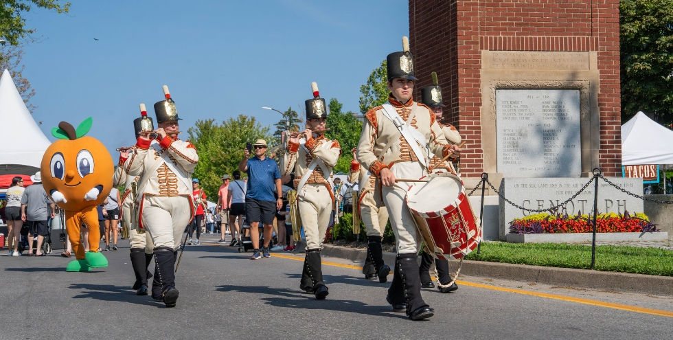 Peach Festival in Niagara-on-the-Lake. Marching band in white uniforms and the Percy the Peach mascot parade past a brick cenotaph monument on a sunny street.
