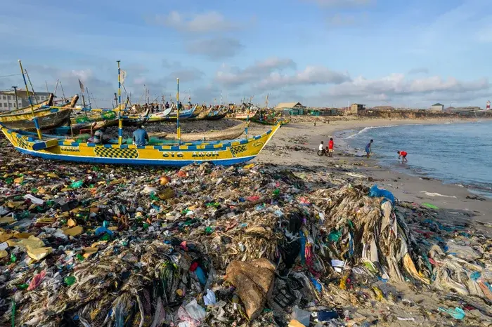 Beached colorful boats on a trash-covered shoreline, with people in the distance by the sea. The sky is clear and blue, creating a stark contrast.
