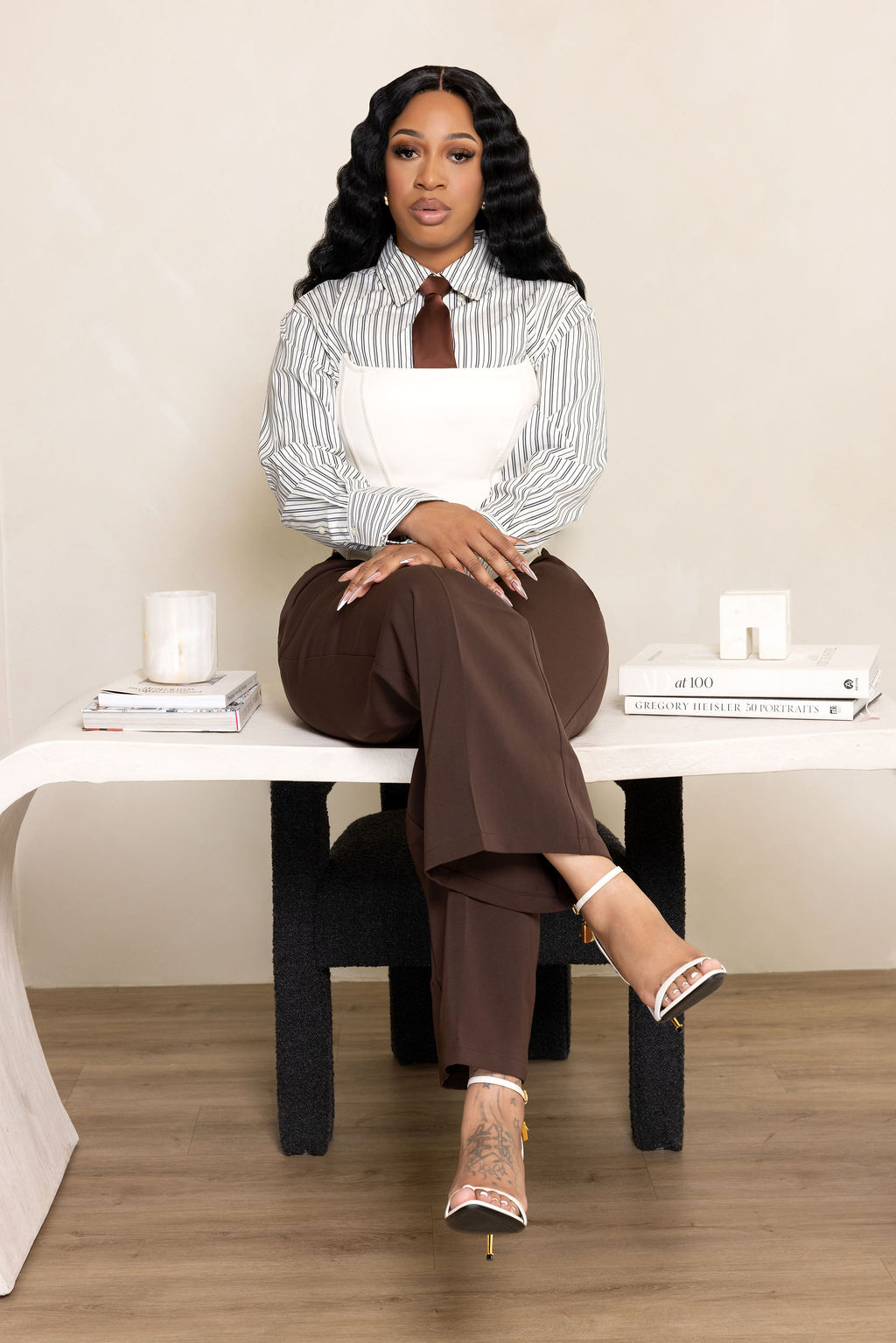 Shae Jordan, founder of Lux Business Firm, seated on a white console in a striped shirt and brown trousers