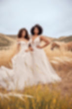 Two woman wearing lace a-line bridal gowns while standing in a wheat field with a golden brown horse in the backround