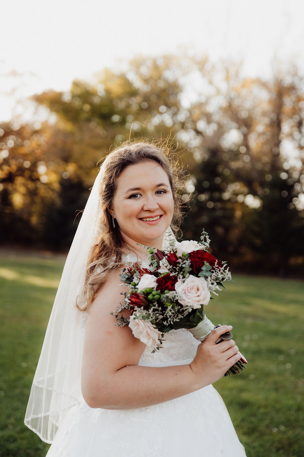 a bride holds her bouquet of flowers up near her face as she smiles while walking outside on a green lawn