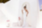 A woman wearing a halter neckline clean cut fitted bridal gown standing in front of and all white stone archway and staircase outside