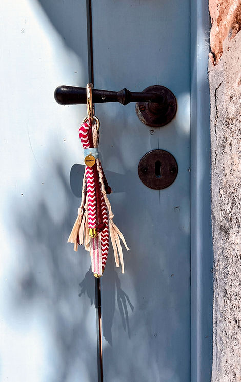 Handgefertigter Cala Bag Charm von SOOOWHAT mit roten Zickzack-Bändern, Bastdetails und goldenen Akzenten – fotografiert an antikem Türgriff