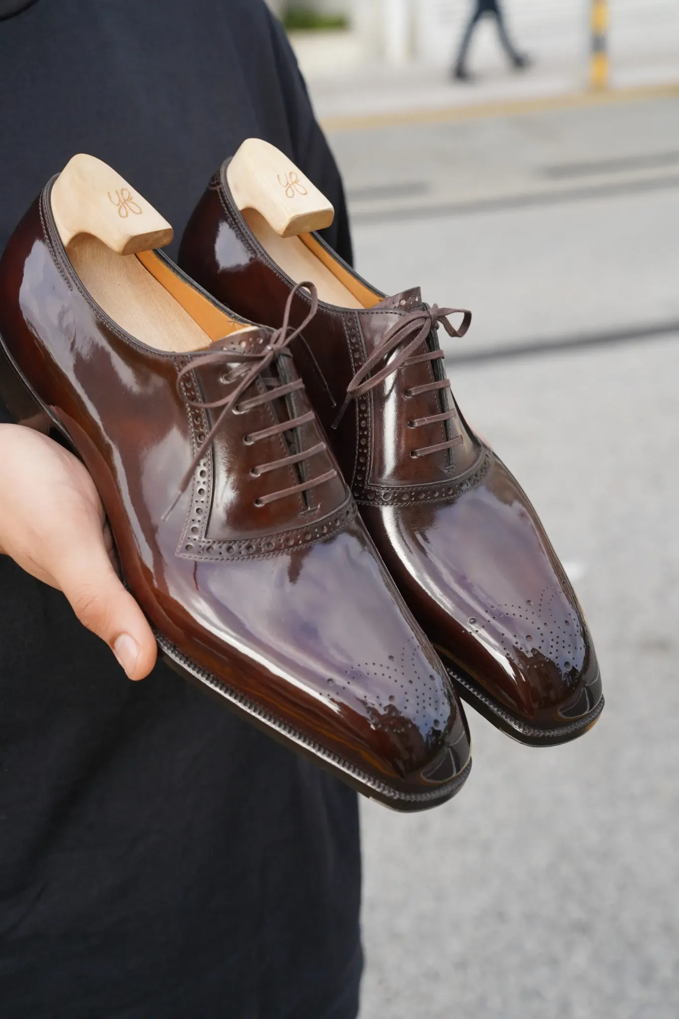 A pair of highly polished brown leather Oxford shoes with wooden shoe trees, held by hand