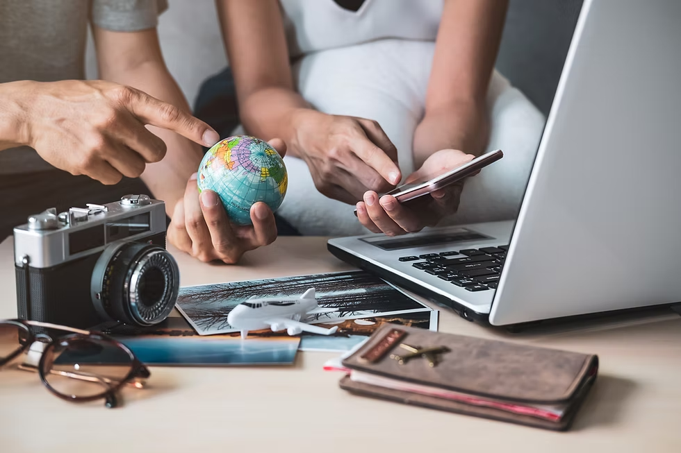Hands holding a globe and smartphone near a laptop, camera, sunglasses, toy plane, and passport, suggesting travel plans.