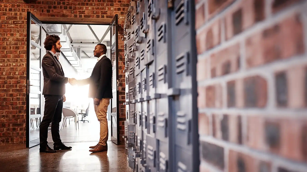 Two men in suits shake hands in a modern office hallway with brick walls and lockers, exuding a professional atmosphere.