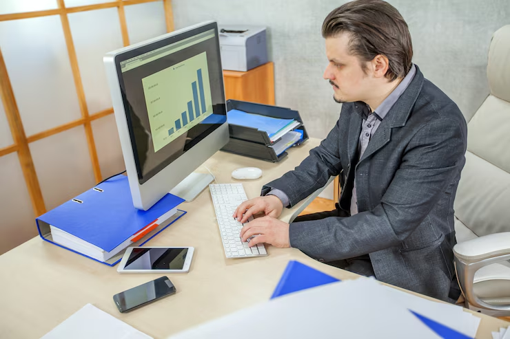 Man in suit analyzes bar graph on desktop monitor; office setting with folders, smartphone, and pens. Focused and professional mood.