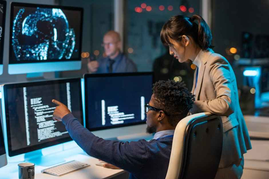 Two people in an office, focusing on computer screens displaying code and a digital globe. Night city lights visible through a window.