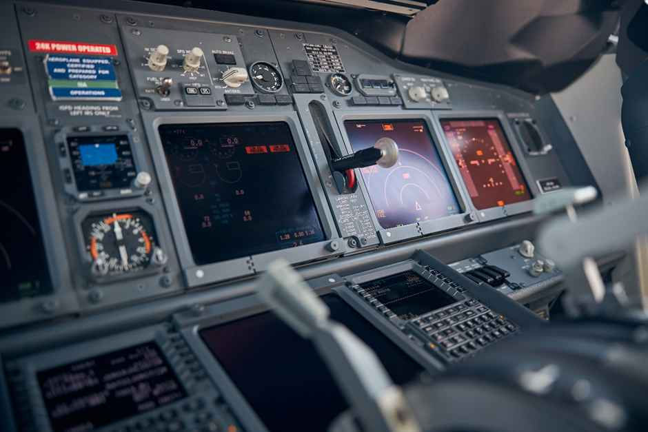 Aircraft cockpit with lit control panels, monitors displaying flight data, and numerous switches and dials. Warm glow on screens.