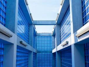 Blue glass walls form a geometric, open-roof corridor. Bright sunlight illuminates the space, creating a modern, serene atmosphere.