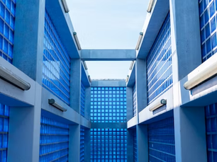 Blue glass walls form a geometric, open-roof corridor. Bright sunlight illuminates the space, creating a modern, serene atmosphere.
