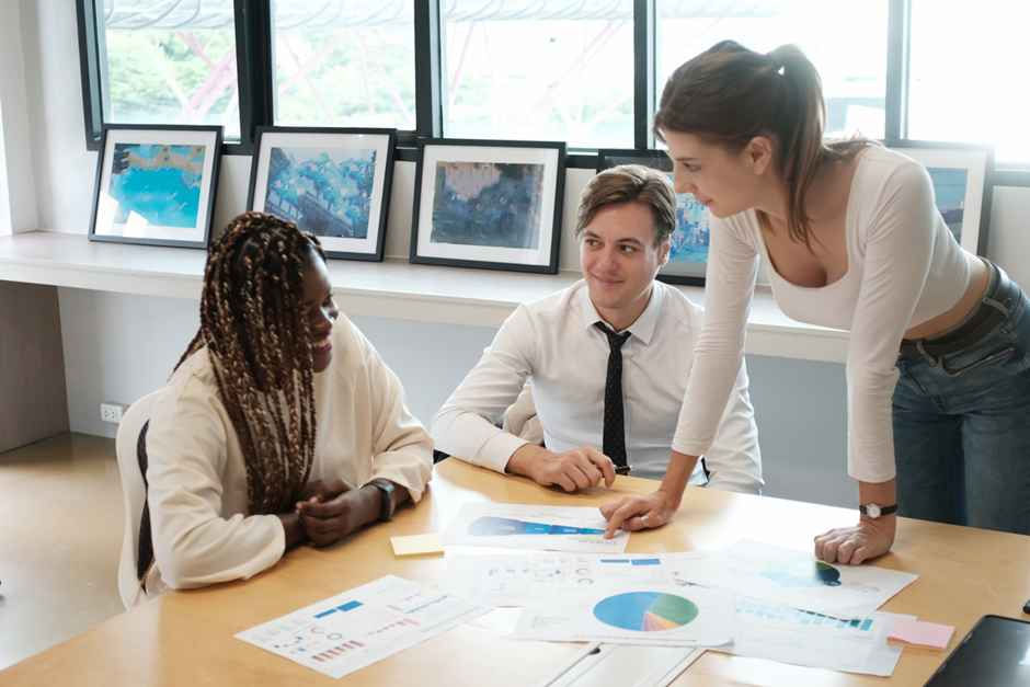 Three people discussing charts at a table in an office with framed art on the windowsill. The atmosphere is collaborative and focused.