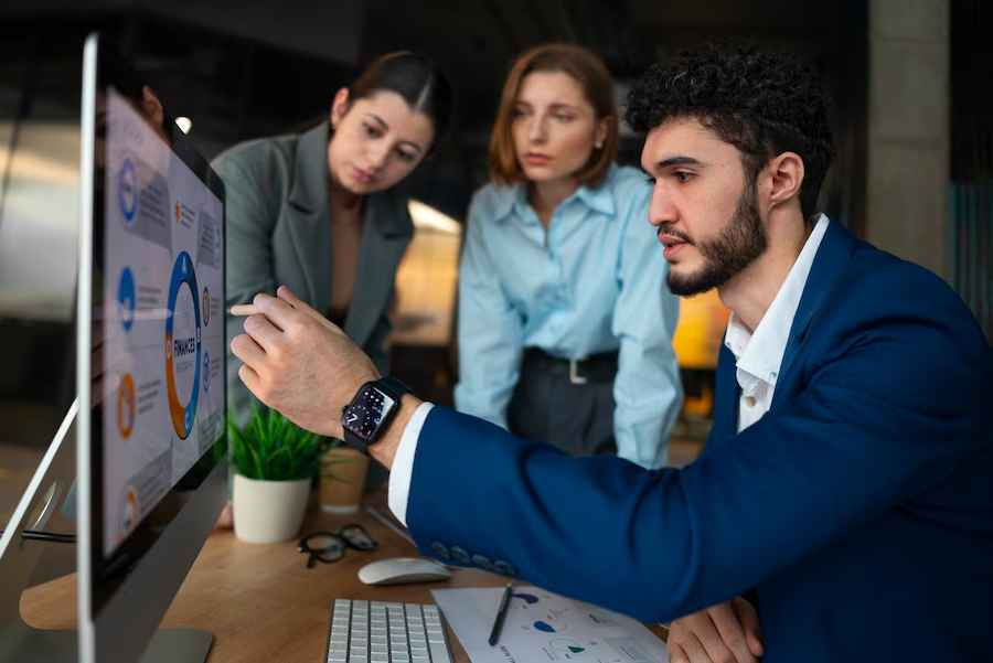 Three people focus on a computer screen showing a circular chart. One points at the screen. Office setting, analytical mood.