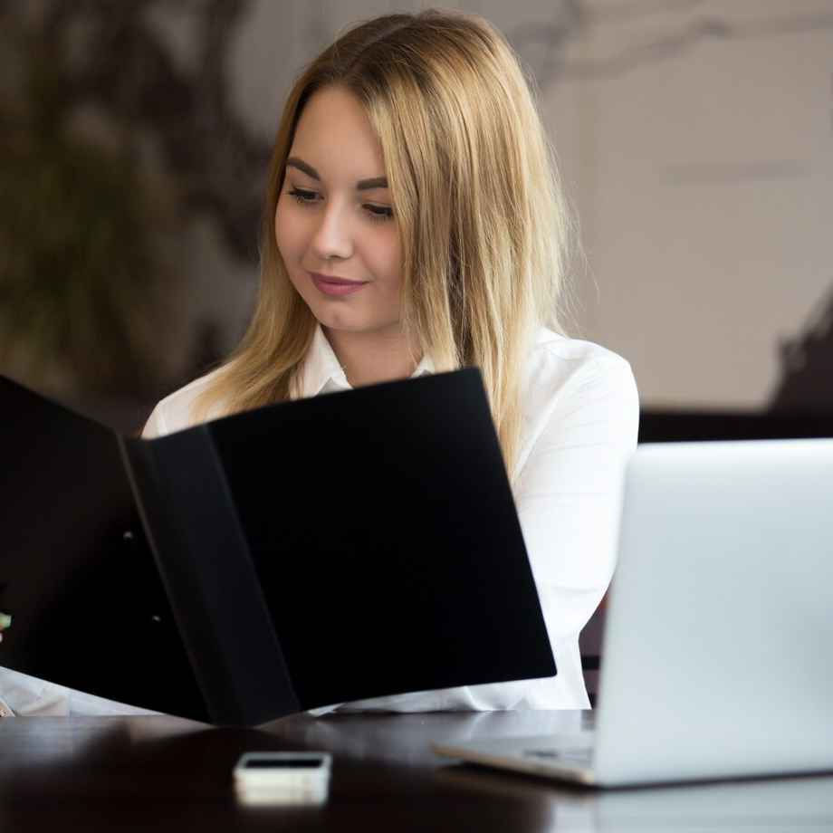 Woman reading a black folder at a desk with a laptop and phone. She's wearing white, and the mood is focused in an office setting.