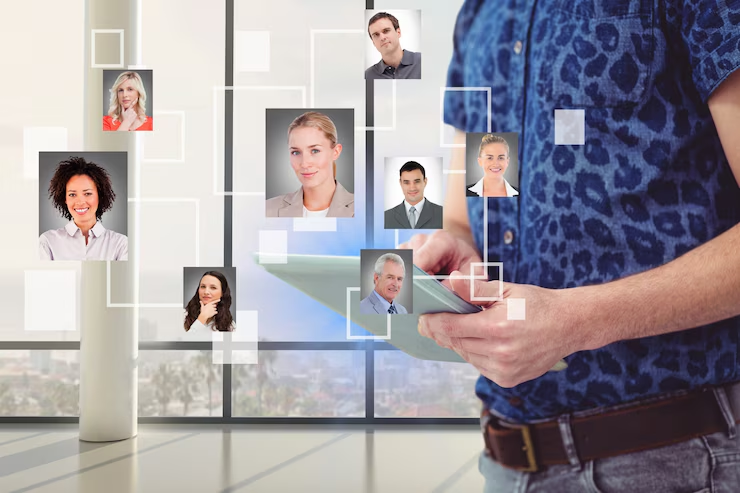 Man in blue shirt holds a tablet in front of a window. Floating portraits of people connected by lines suggest a digital network.