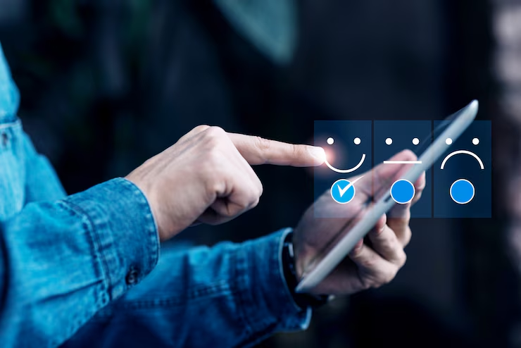 Hands in a denim jacket tap a tablet screen showing smiley faces for feedback. The selected face is happy. Dark blurred background.