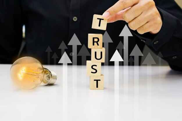 Person stacking wooden blocks spelling "TRUST" beside a glowing bulb on a white table. Background has upward arrows. Business concept.