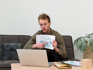 Man in glasses sits on a sofa, pointing to a chart while on a laptop. Papers and a notebook are on the table. Calm, indoor setting.