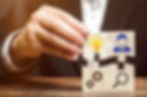 A hand in a suit holds puzzle pieces with icons of a light bulb, person, gears, and magnifying glass. Wood surface and blurred brown background.
