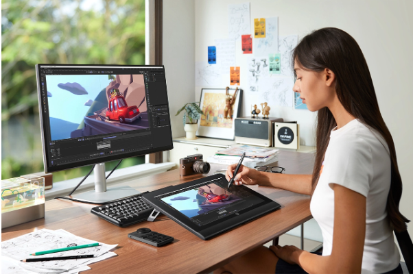 Woman using a stylus to draw on a tablet connected to a monitor displaying animation. Desktop with keyboard, camera, art in bright room.
