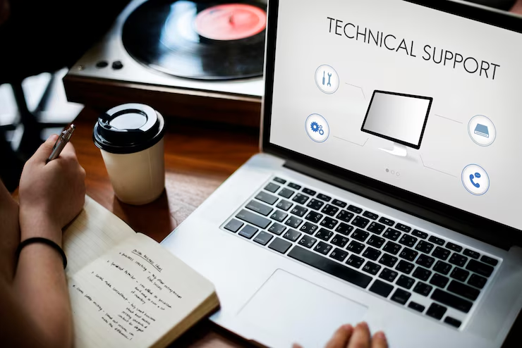Laptop showing "TECHNICAL SUPPORT," person taking notes in notebook, coffee cup nearby, vinyl record in background, calm work setting.