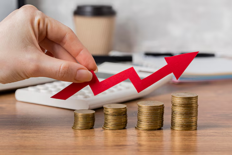 Hand holding a red zigzag arrow above stacks of coins on a desk, symbolizing financial growth. Calculator and notebook in background.