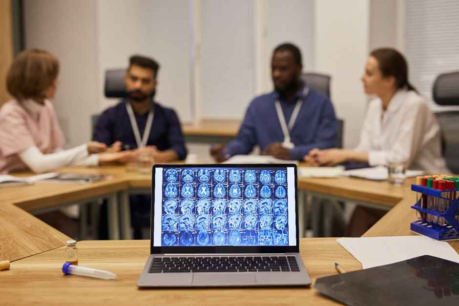 Laptop with brain scans on screen in foreground. Four people in a meeting in the background, discussing in an office setting.