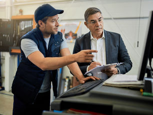Man in cap points at screen while another in a suit holds a clipboard. They're in a workshop with tools in the background.