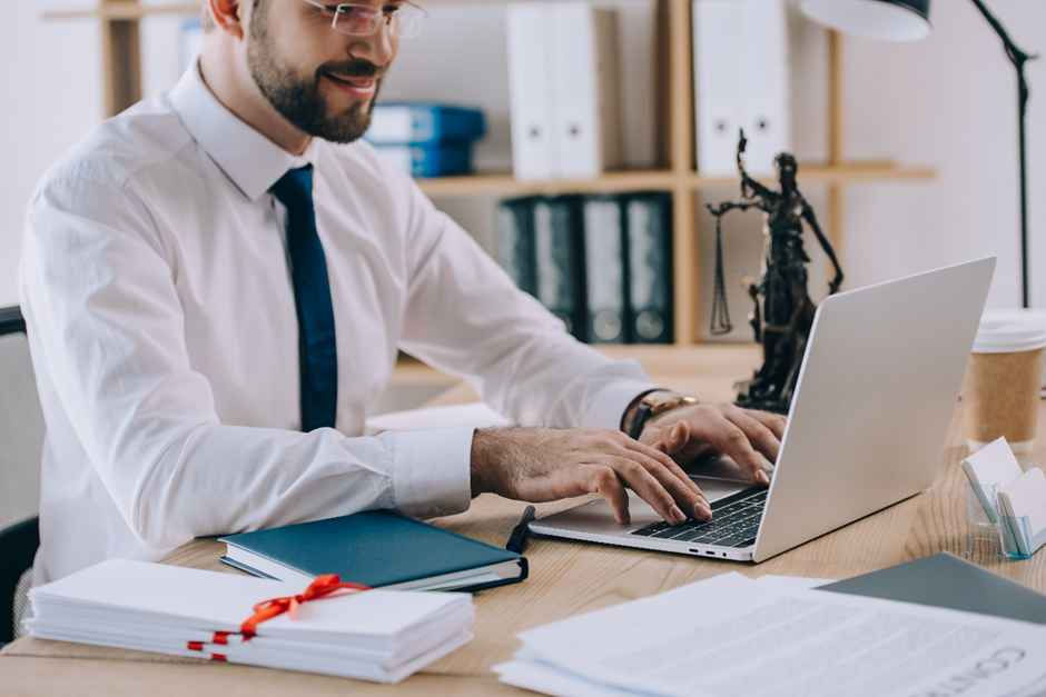 Man in a white shirt typing on a laptop at a desk with books, papers, and a justice statue. Background shelves with files. Professional mood.