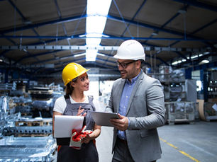 Two people in hard hats chat in a warehouse. One holds documents, the other a tablet. Steel racks in the background. Bright, professional mood.