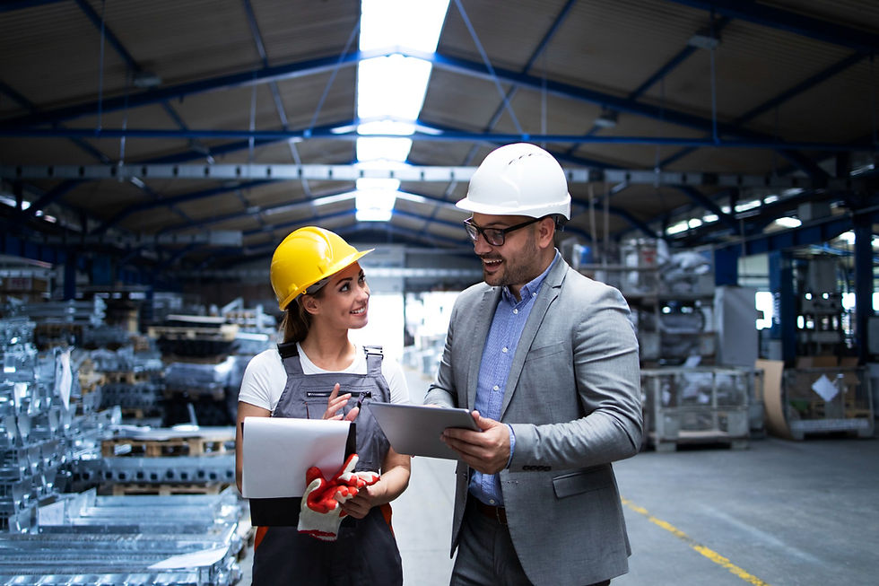 Two people in hard hats chat in a warehouse. One holds documents, the other a tablet. Steel racks in the background. Bright, professional mood.