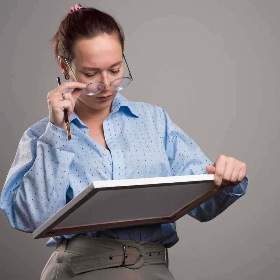 A woman in a blue shirt examines a painting canvas, holding a pencil and adjusting her glasses, in a neutral, gray background setting.