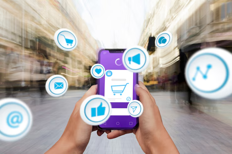 Hands hold a purple smartphone displaying a shopping cart app. Surrounding icons include email and like symbols, blurred city street background.