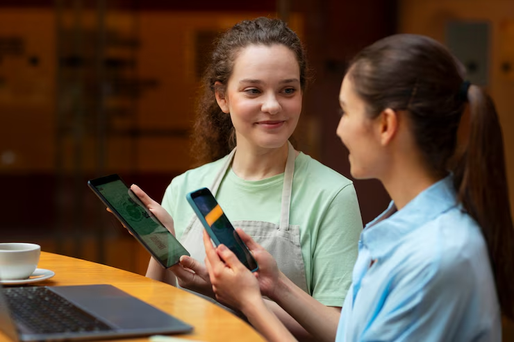 Two women at a cafe, smiling and holding a tablet and smartphone, one in an apron. A cup on the table, blurred background. Warm atmosphere.