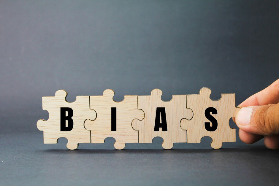 Wooden puzzle pieces spelling "BIAS" held by a hand on a gray background, suggesting a concept or theme.