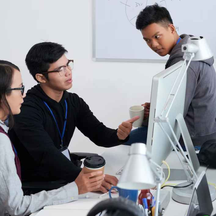 Three people in an office discuss work at a computer. Two hold coffee cups. A whiteboard is in the background. The mood is focused.
