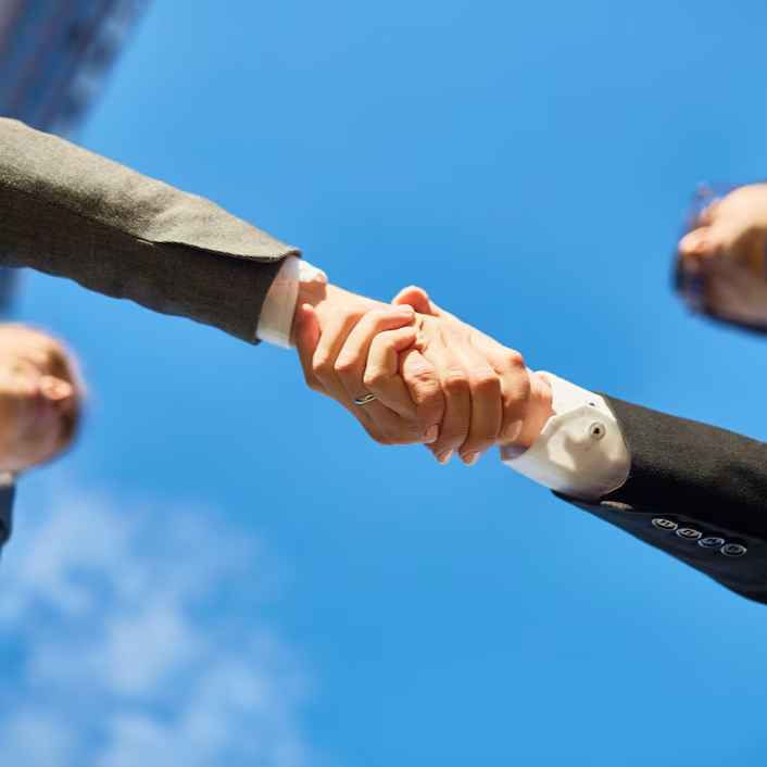Two people in suits shaking hands under a clear blue sky, viewed from below. Partially visible faces suggest collaboration and agreement.