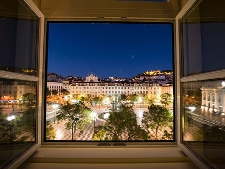Vista noturna de Lisboa através de uma janela aberta. Praça iluminada com edifícios históricos e árvores. Céu azul profundo ao fundo.