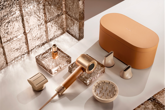 Itens de beleza dourados em uma superfície clara: secador, bico, difusor e frasco, em cena elegante com fundo texturizado prateado.
