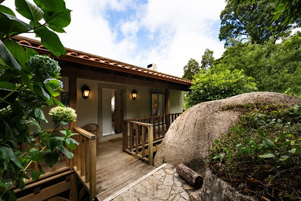 Casa rústica de madeira com varanda, cercada por vegetação verde e rocha. Céu azul ao fundo. Ambiente tranquilo e acolhedor.