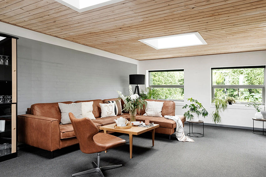 Sala de estar aconchegante com sofá marrom, mesa de madeira, plantas e janelas grandes. Luminária preta e luz natural destacam o ambiente.