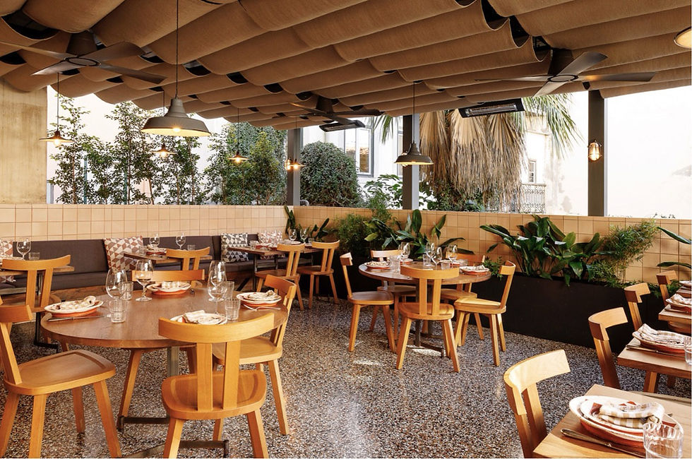 Restaurante elegante com mesas de madeira e louças arrumadas. Teto de tecido ondulado, plantas e luzes pendentes. Ambiente acolhedor.