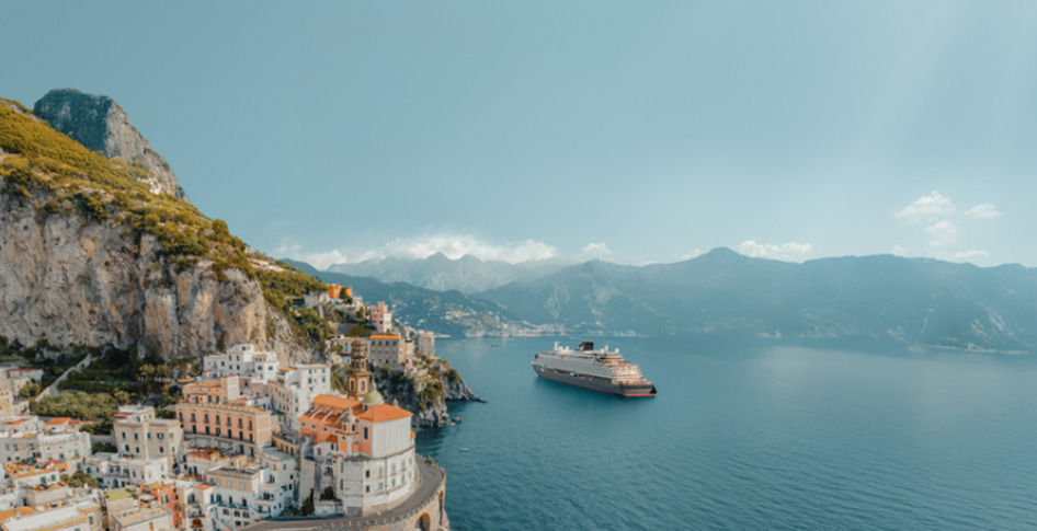 Costa ensolarada de Amalfi com casas coloridas ao lado de penhasco. Cruzeiro navega pelo mar azul sob céu claro, transmitindo tranquilidade.