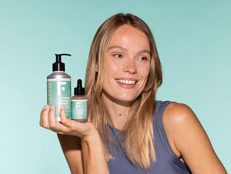 Mulher sorrindo segura produtos de cabelo em fundo azul claro. Ela veste blusa cinza. Embalagens verdes com texto visÃvel. Emocional alegre.