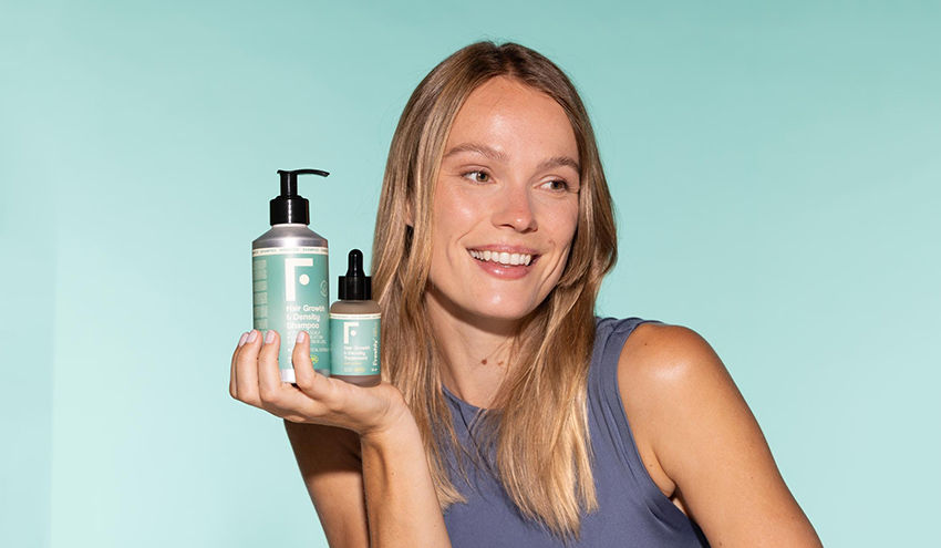 Mulher sorrindo segura produtos de cabelo em fundo azul claro. Ela veste blusa cinza. Embalagens verdes com texto visível. Emocional alegre.