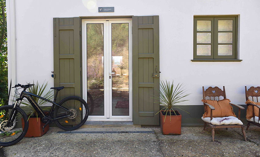 Bicicleta encostada em parede branca com porta de vidro e janelas. Vasos de plantas e cadeiras de madeira com almofadas laranja ao lado.