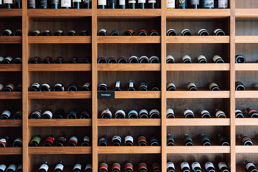 Garrafas de vinho em estantes de madeira em uma adega. As prateleiras estão organizadas e algumas garrafas são coloridas. Ambiente tranquilo. Enoteca do Octant